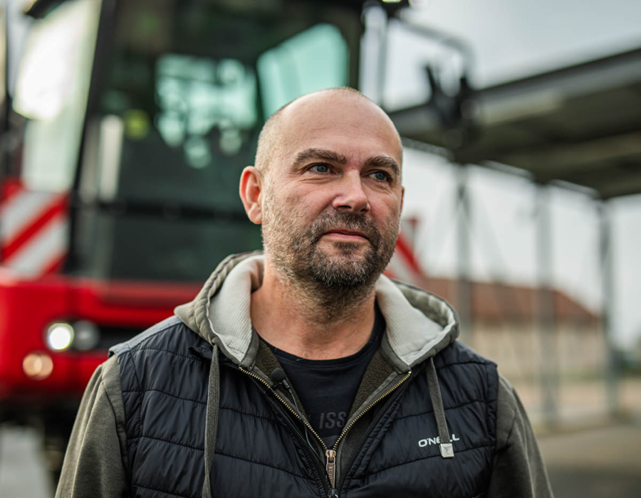 A bald man stands confidently in front of a bright red tractor, showcasing a rural setting and agricultural machinery.