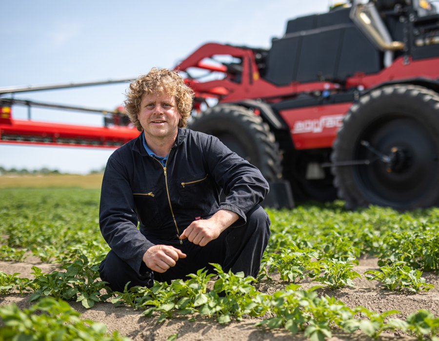 A man kneels in front of an Agrifac sprayer in a field, inspecting the field surrounded by open land and greenery.