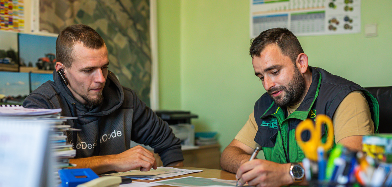 Two men seated at a table, engaged in discussion with papers and pens in front of them.