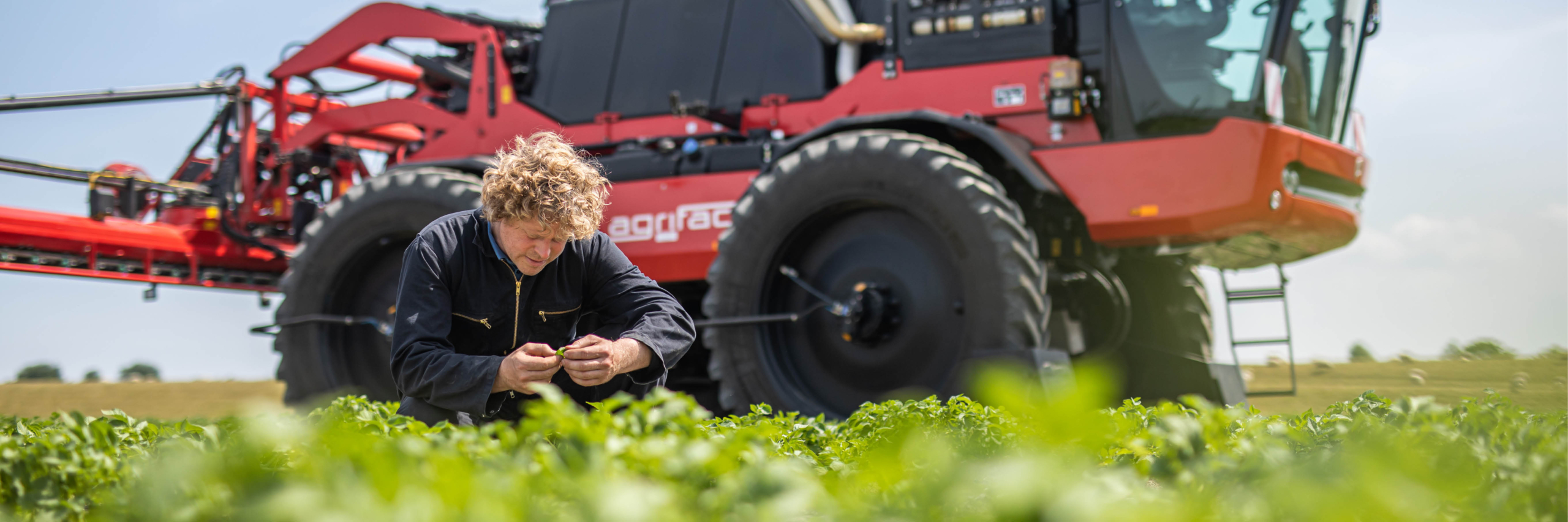 A man kneels in the field besides his Agrifac sprayer, engaged in an outdoor task, surrounded by a lush green landscape.