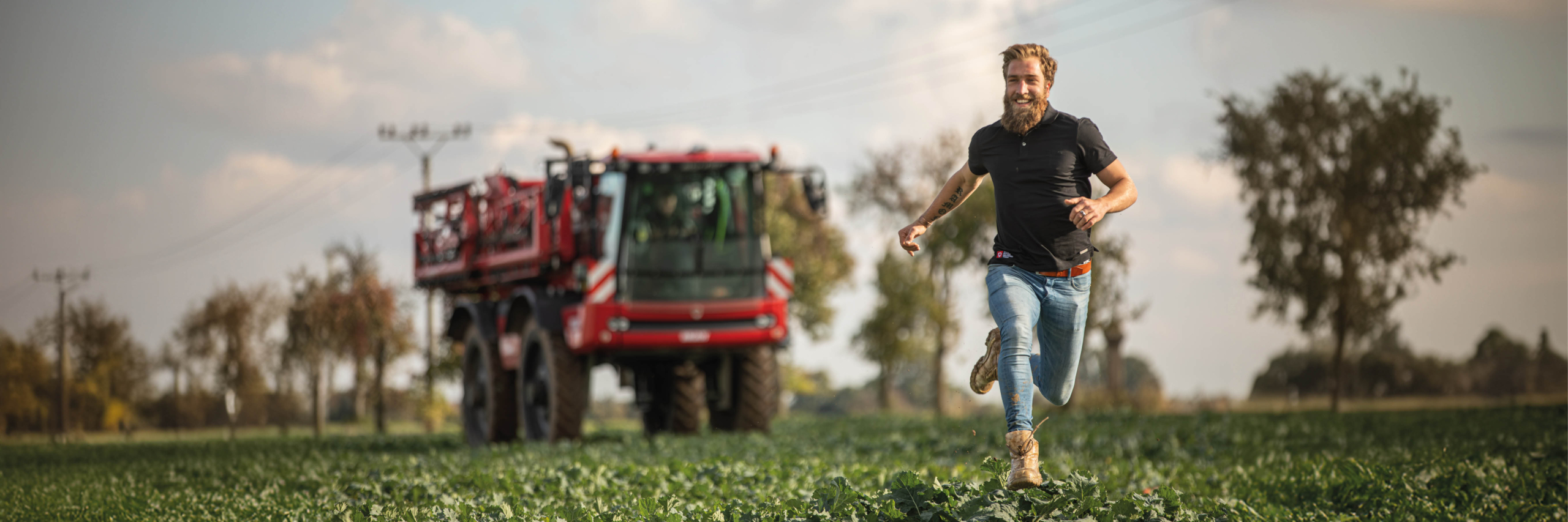 A man runs energetically through a field, with an Agrifac sprayer visible in the background, showcasing rural life and activity.